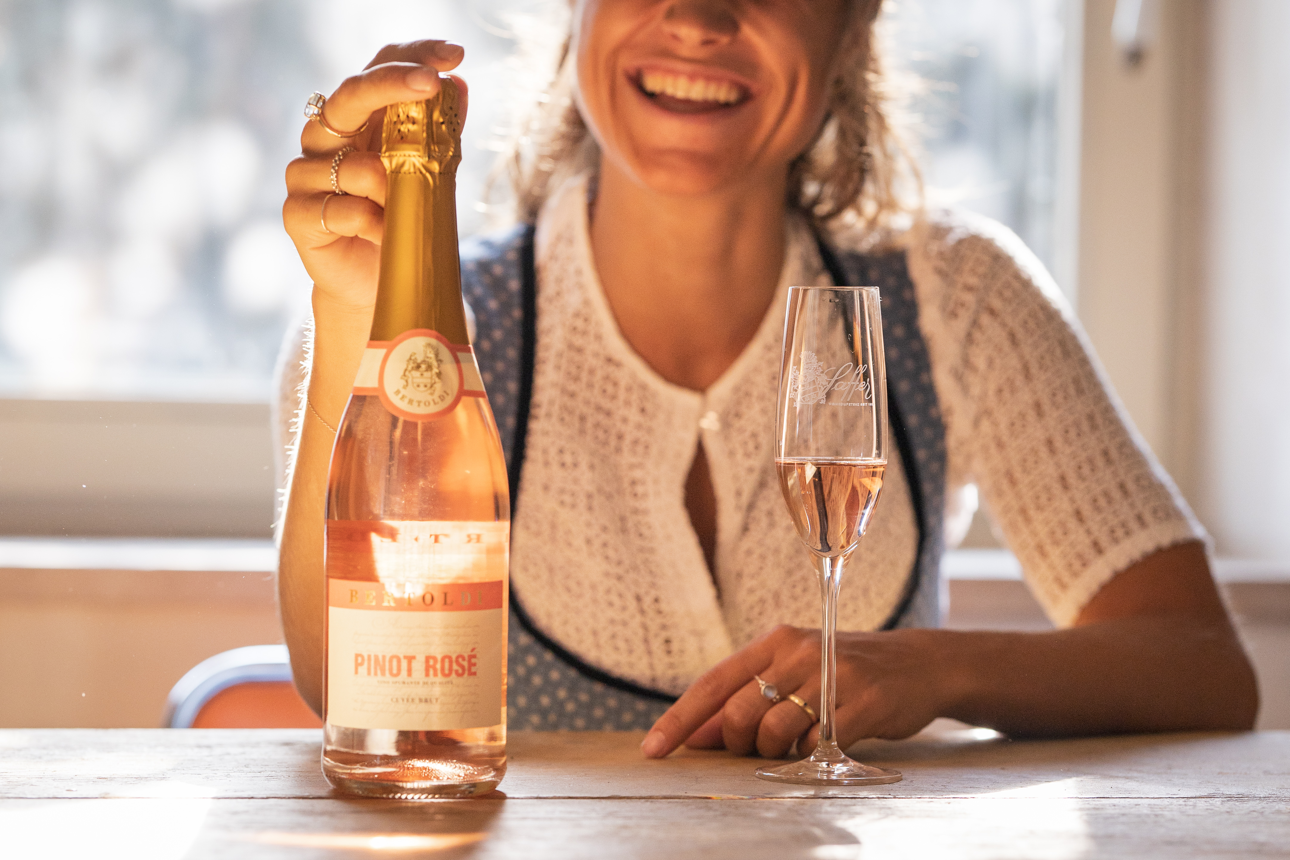 Fröhliche Frau im Dirndl sitzt an einem Tisch mit einem Glas Rosé-Sekt in der Hand und präsentiert eine Flasche Pinot Rosé, Sonnenlicht fällt durch das Fenster.