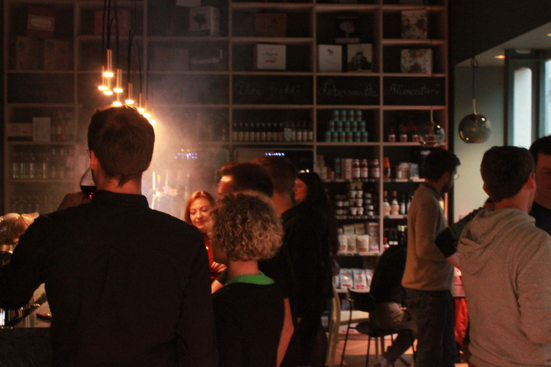 Abendliches Get-together in einer Weinhandlung mit vielen Gästen, im Vordergrund eine Personengruppe bei Gesprächen, im Hintergrund ein gut bestücktes Regal mit Wein und Feinkost.