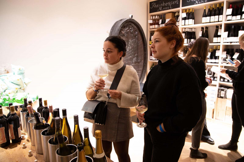Zwei Frauen bei einer Weinverkostung in einer Vinothek, mit gefüllten Weingläsern in der Hand, umgeben von gekühlten Weinflaschen und vollen Regalen im Hintergrund.