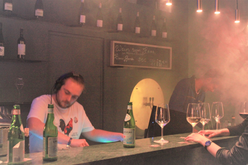 DJ legt bei ‚Bubbles & Beats‘ in Saffer’s WinzerWelt inmitten von Nebel und Partystimmung auf, mit Weingläsern und Flaschen auf der Theke.