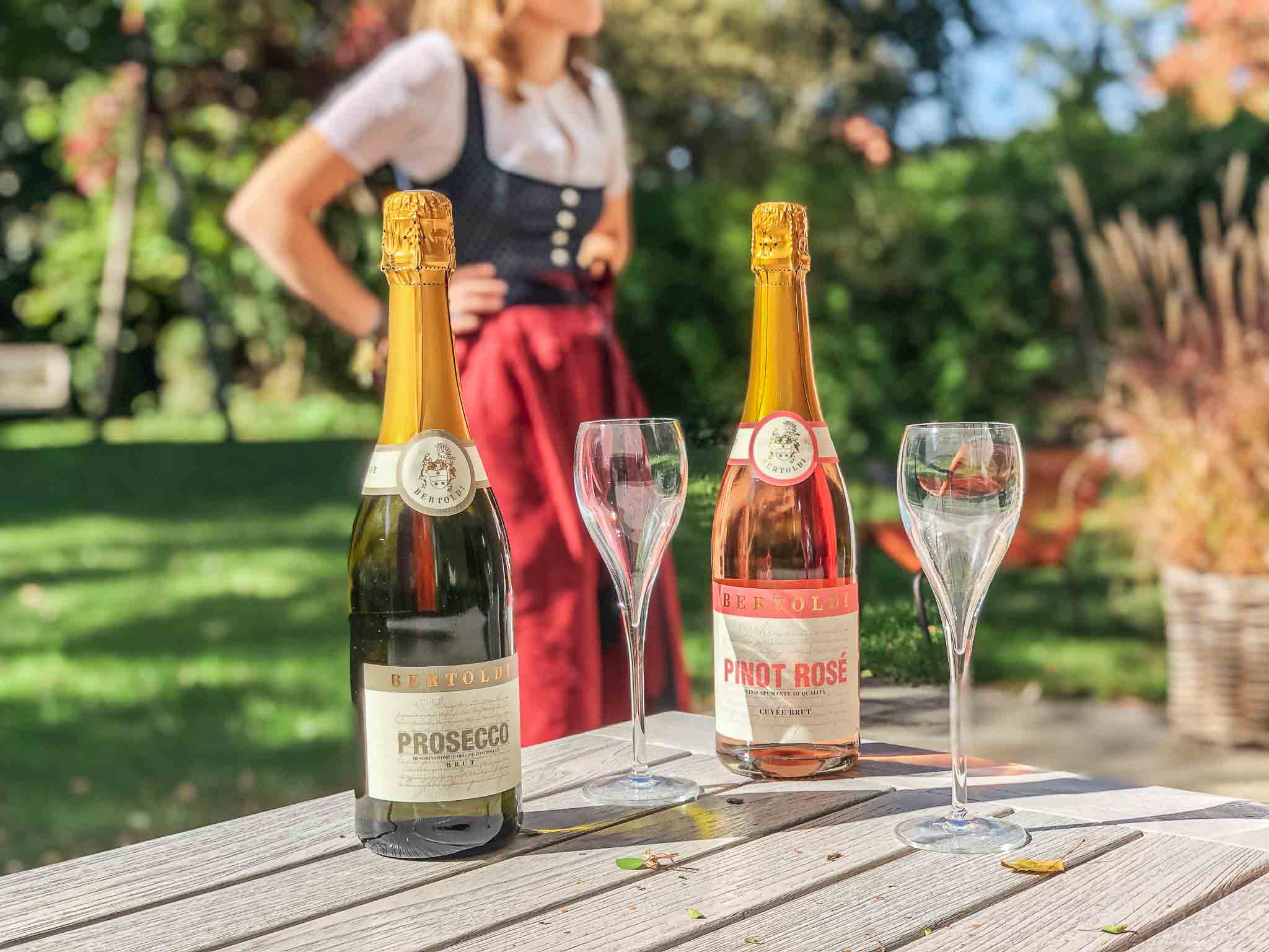 Zwei Flaschen Schaumwein – Prosecco und Pinot Rosé – mit Sektgläsern auf einem Holztisch im Garten, im Hintergrund eine Frau in Dirndlkleidung bei sonnigem Herbstwetter.
